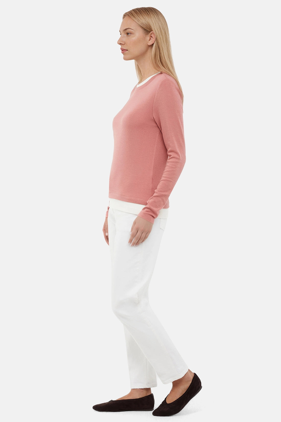 Woman wearing a pink sweater with white layered hem and white pants on a light gray background.