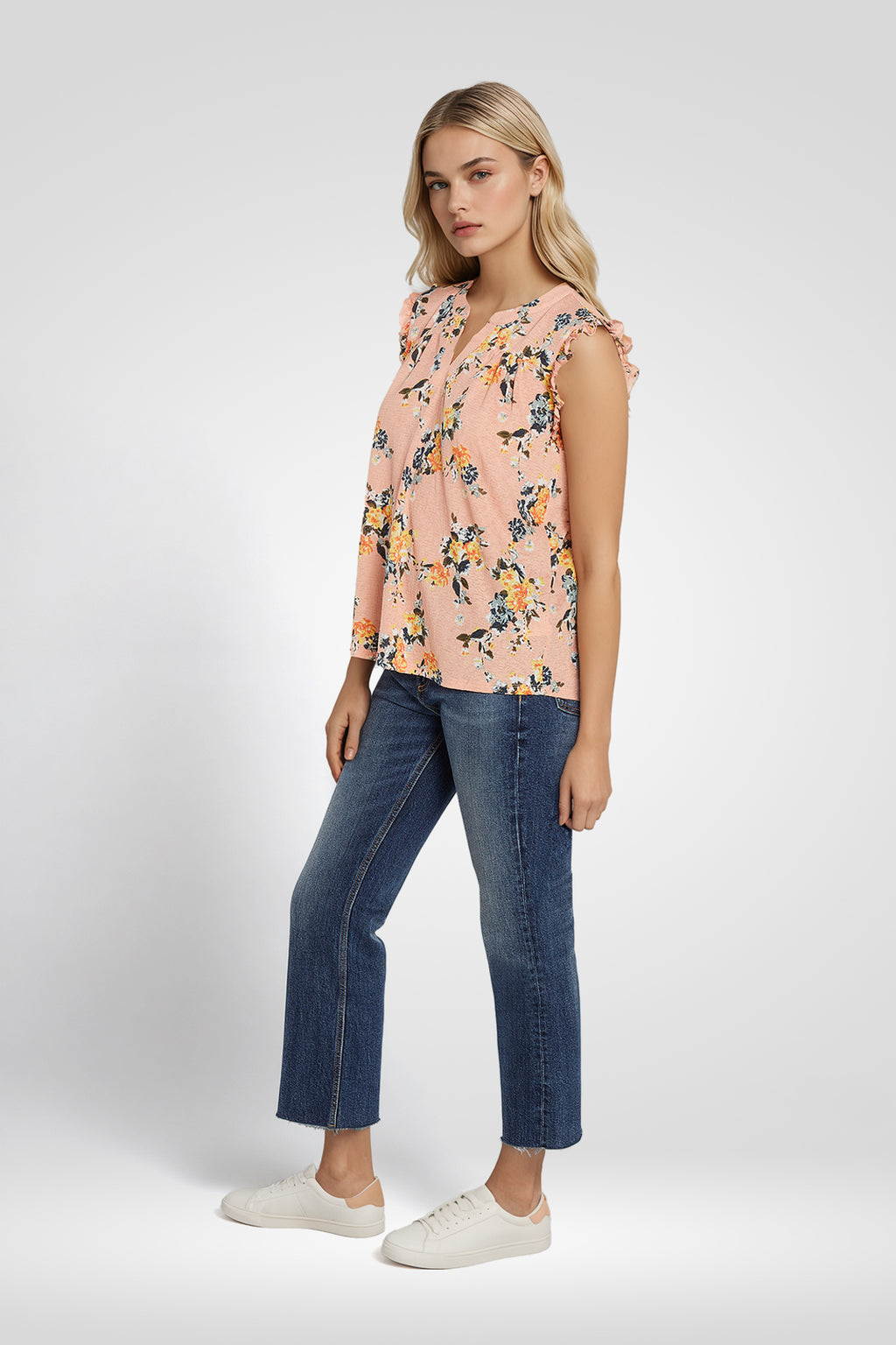 Woman wearing a sleeveless floral blouse and blue jeans with a plain background. coral-pink-yarrow