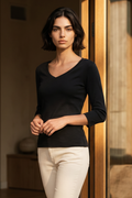 A woman with short dark hair stands indoors wearing a fitted black long-sleeve V-neck top and light cream pants, posed in front of a warm-toned wooden door.