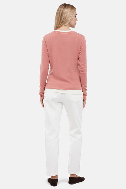 Back of woman wearing a pink sweater with white layered hem and white pants on a light gray background. 