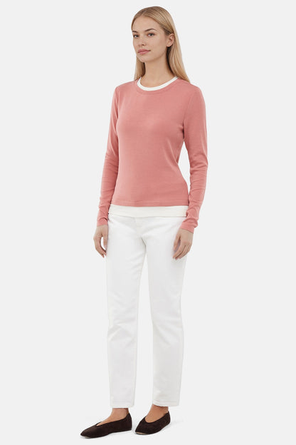 Woman wearing a pink sweater with white layered hem and white pants on a light gray background. 