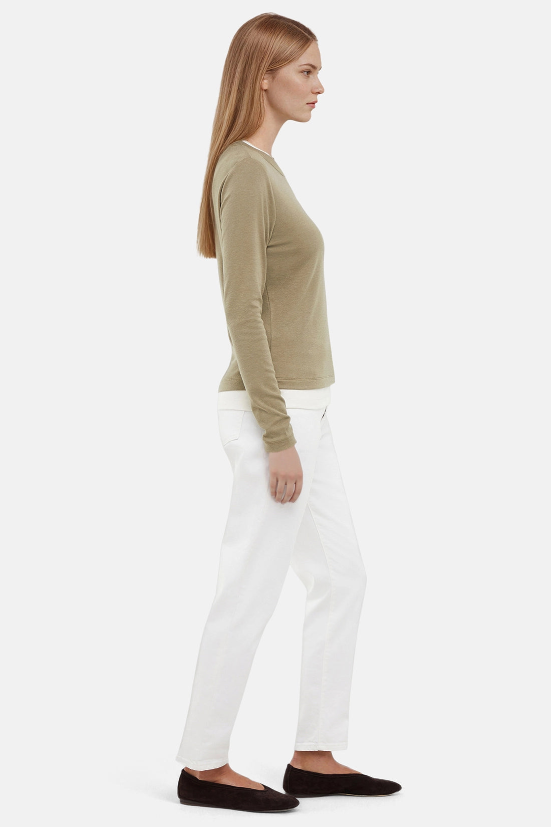 Side view of woman wearing a green sweater with white layered hem and white pants on a light gray background. 
