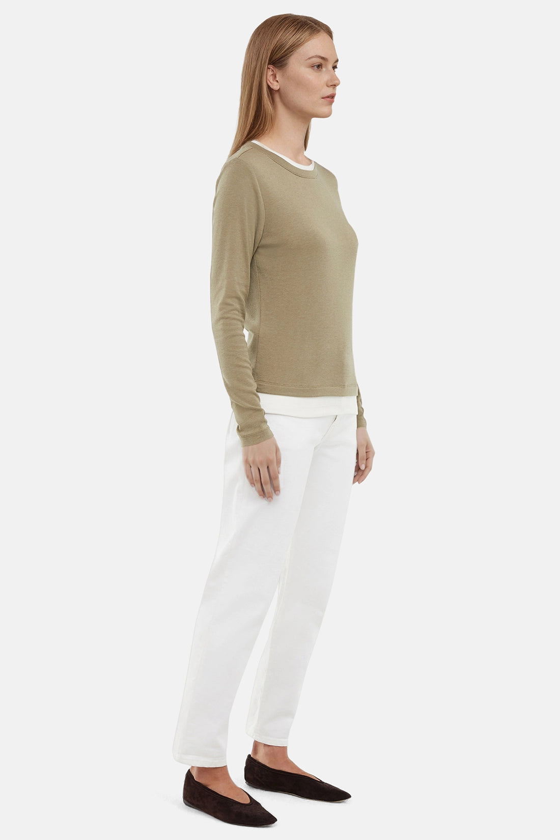 Side view of woman wearing a green sweater with white layered hem and white pants on a light gray background. 