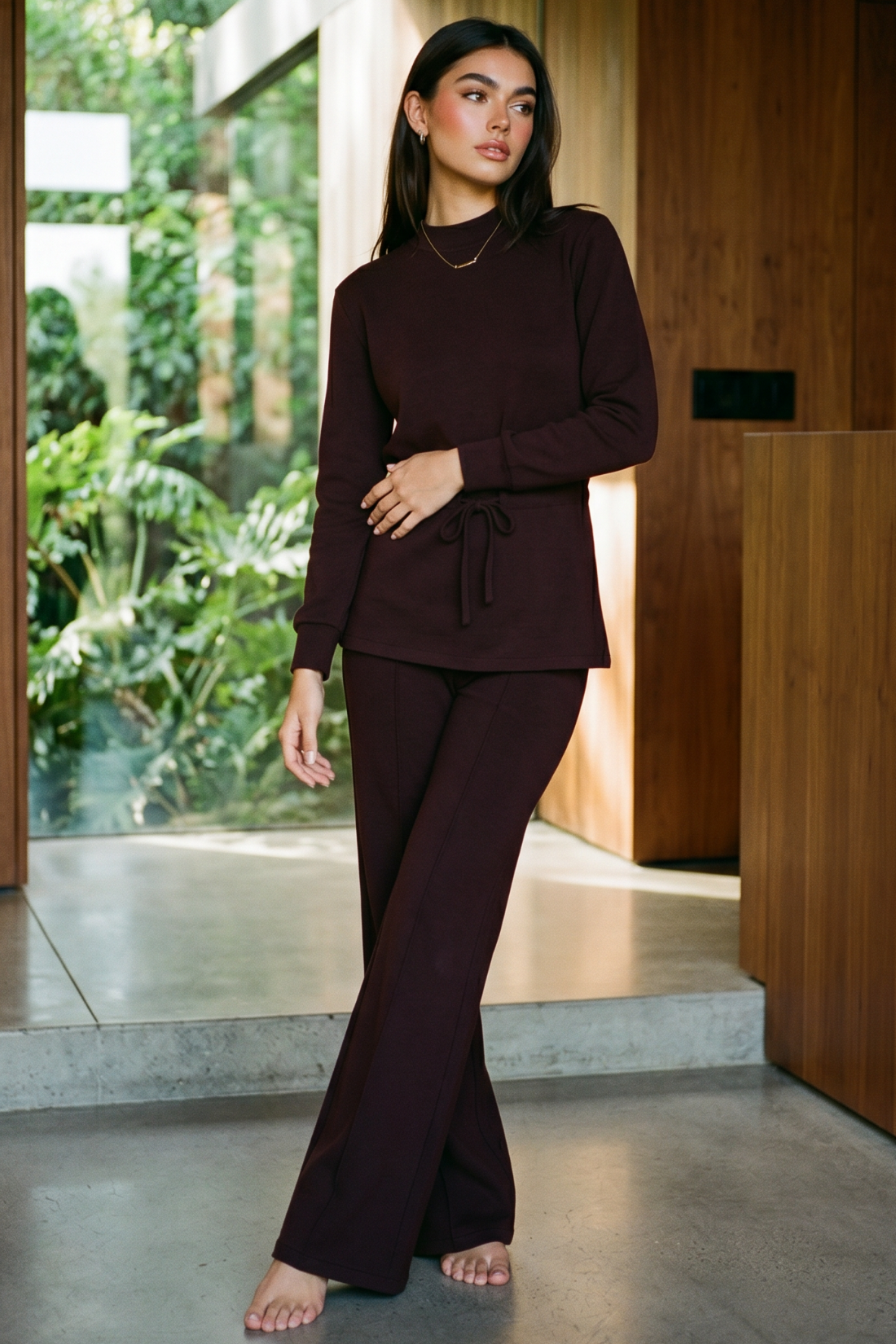 A woman modeling a matching dark plum long-sleeve top and wide-leg pants set in a modern indoor space with large windows and greenery outside. 
