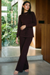 A woman modeling a matching dark plum long-sleeve top and wide-leg pants set in a modern indoor space with large windows and greenery outside. 