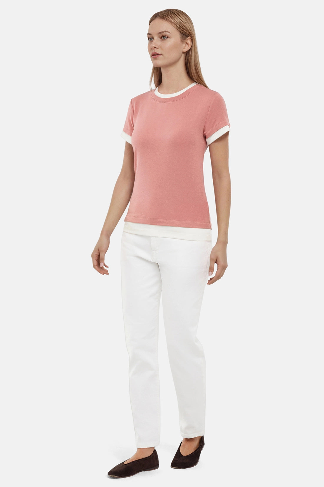 Side view of woman wearing a pink tee with white layered hem and white pants on a light gray background 