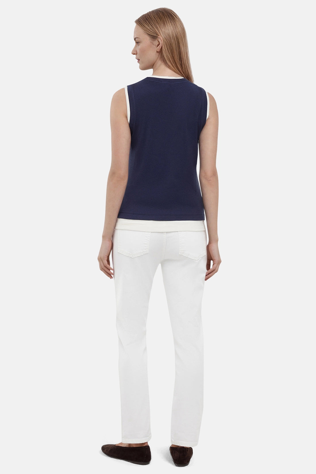Back of woman wearing a navy tank with white layered hem and white pants on a light gray background. 