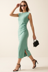 A woman models a sleeveless sage green midi dress with a side slit, styled with black strappy heels, sunglasses, and a small black handbag against a plain beige studio background. 