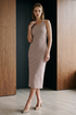A woman models a sleeveless, V-neck midi dress with diagonal stripes in beige and taupe, paired with nude heels, set against a modern interior background with wood paneling and a dark wall. 