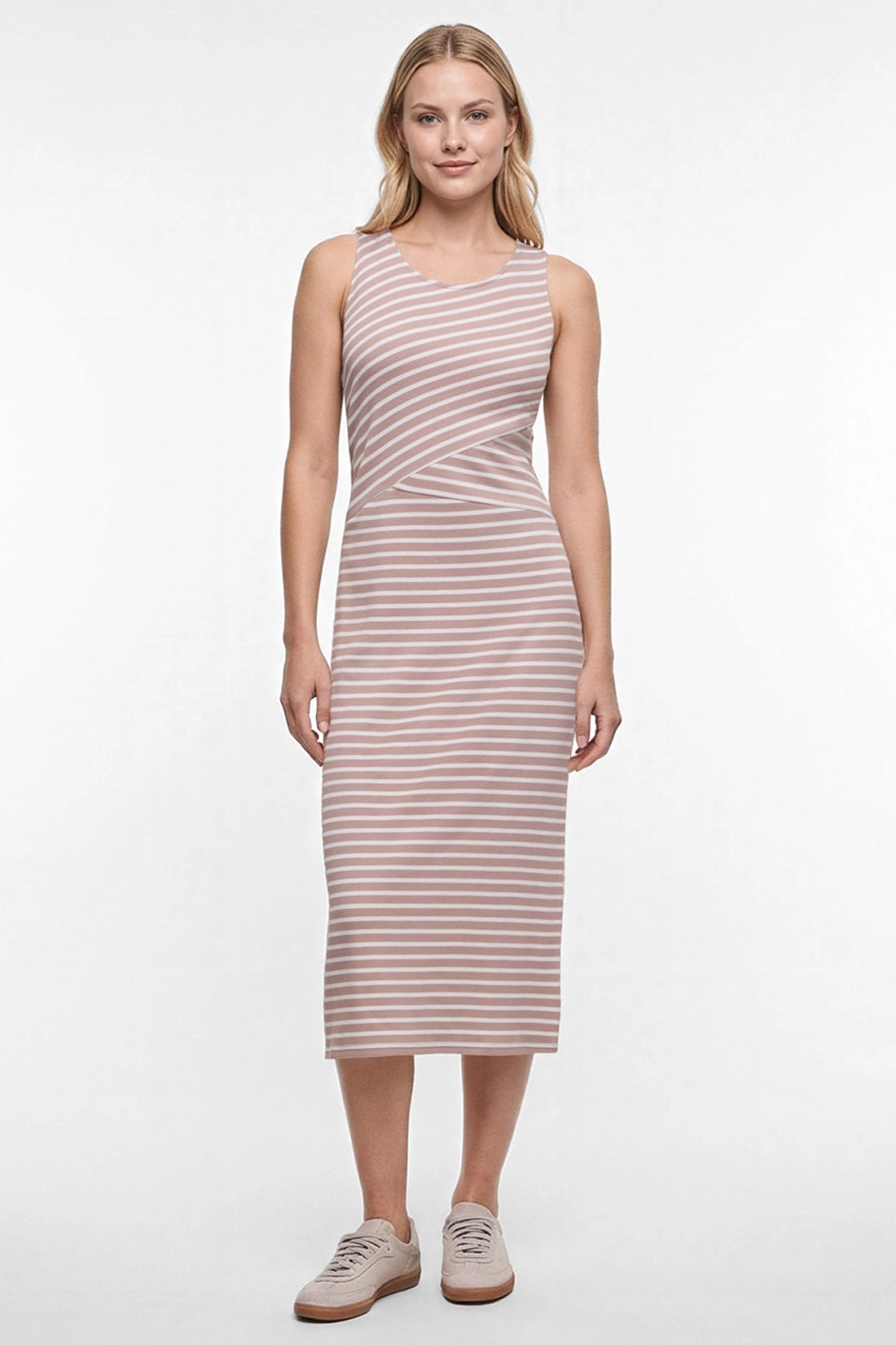 A woman modeling a sleeveless midi dress with horizontal light brown and white stripes, paired with beige sneakers, against a plain white studio background. 