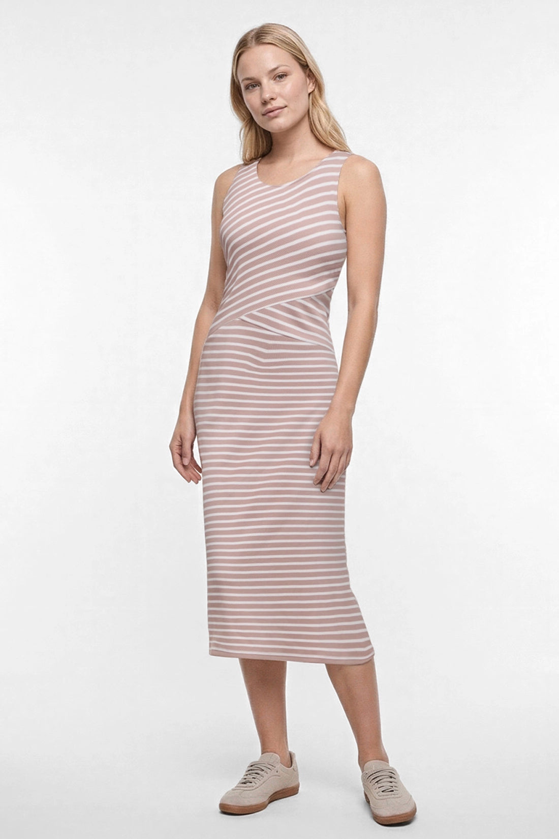 A woman modeling a sleeveless midi dress with horizontal light brown and white stripes, paired with beige sneakers, against a plain white studio background. 