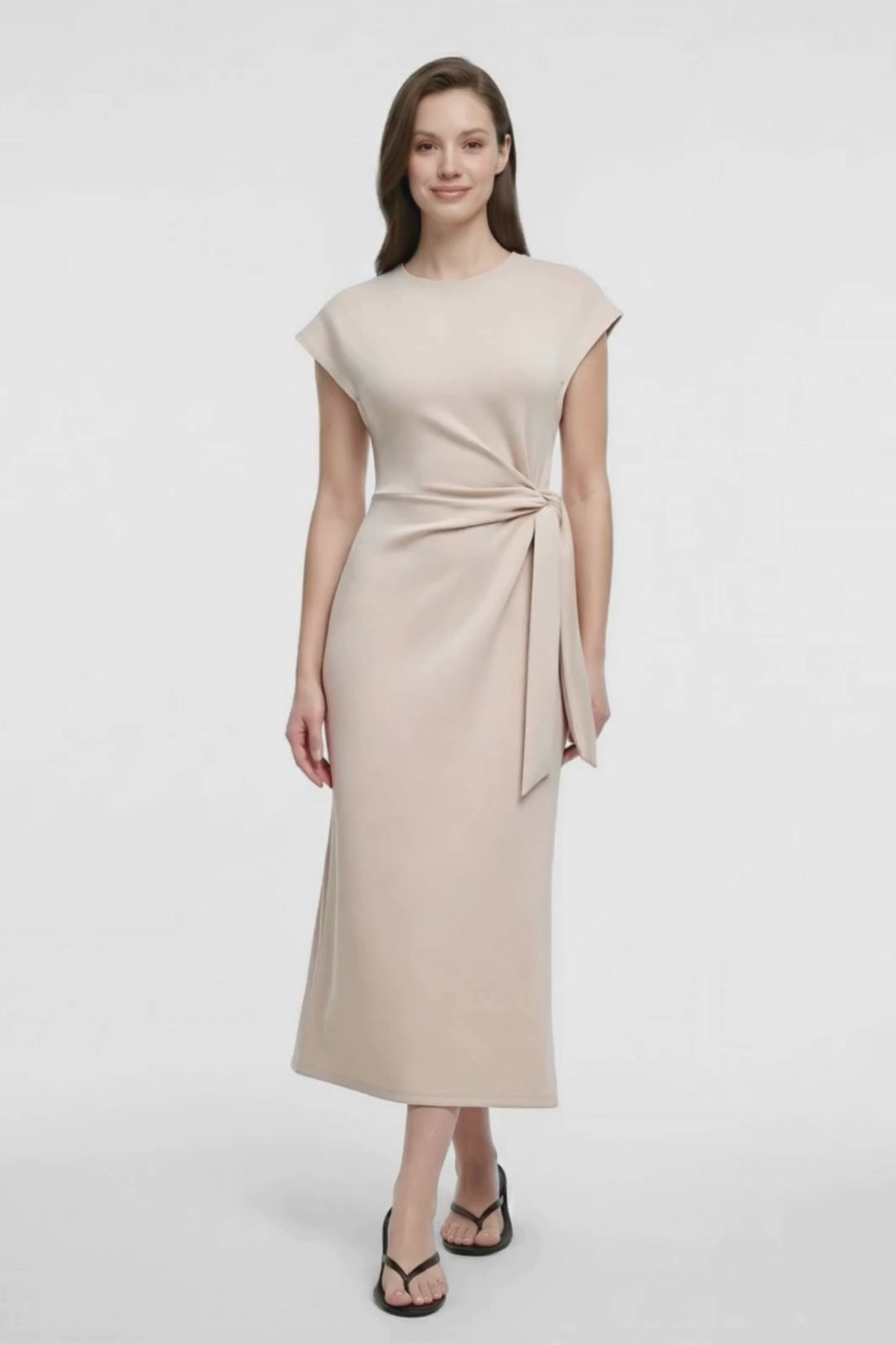 Video view of woman wearing a beige dress with tie detail at waist on a white background. 